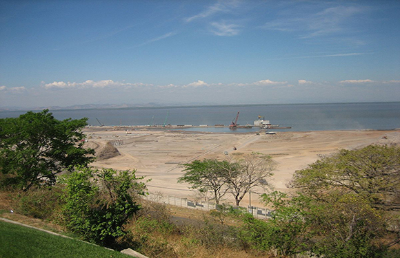 A coastal scene with a sandy beach, calm blue water, clear sky, boats near the shore, and green foliage on the left.