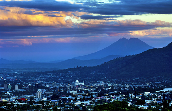 A cityscape during twilight with buildings in the foreground and mountains in the background under a sky with clouds that are lit by the setting sun.