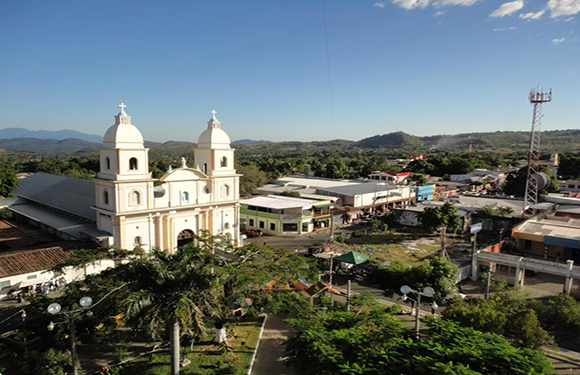 Aerial view of a town with a white church, two bell towers, buildings, greenery, hills, and clear sky.