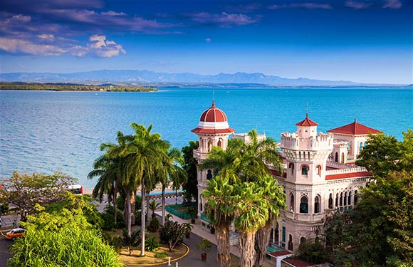 A picturesque coastal scene with a white building with red roofs, palm trees, calm blue sea, distant mountains, and clear sky.