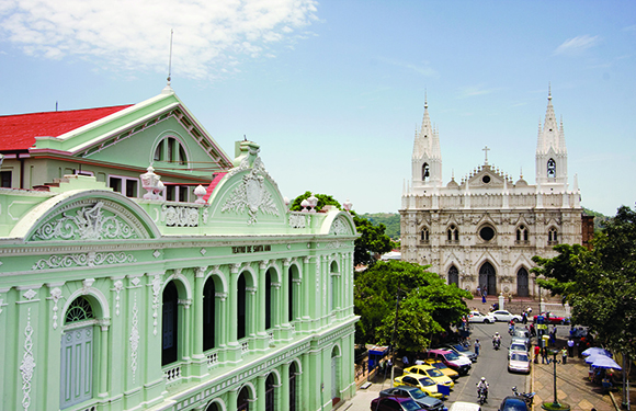 A cityscape with a green historic building, a white cathedral with two spires, a street with cars and people under a clear sky.