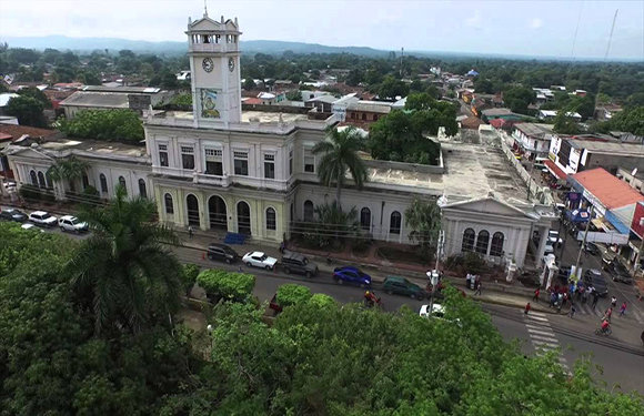 Aerial view of a historic building with a clock tower, surrounded by trees and streets with a few cars, under a cloudy sky.