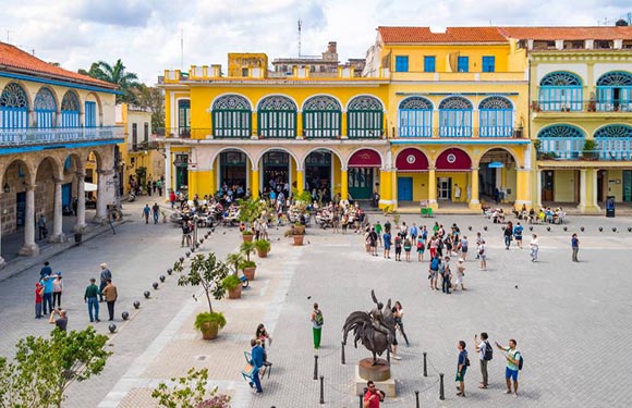 A lively town square with colonial buildings, people, and a central statue under a partly cloudy sky.