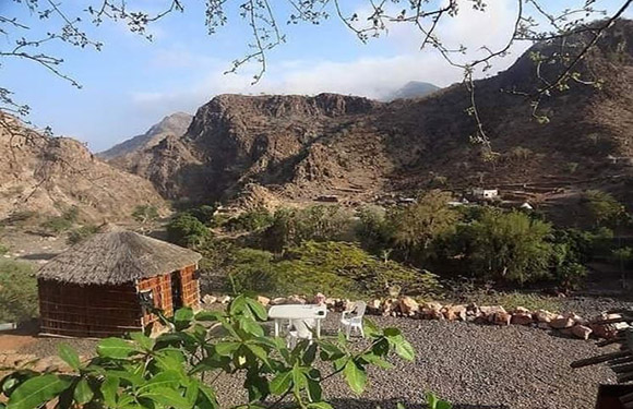 A rural scene featuring a hut, goats, mountains, green shrubs, and a partly cloudy sky.