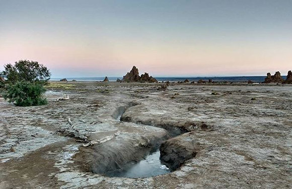 A twilight coastal scene with distant rock formations, a stream to the sea, and a muddy, sparsely vegetated foreground.