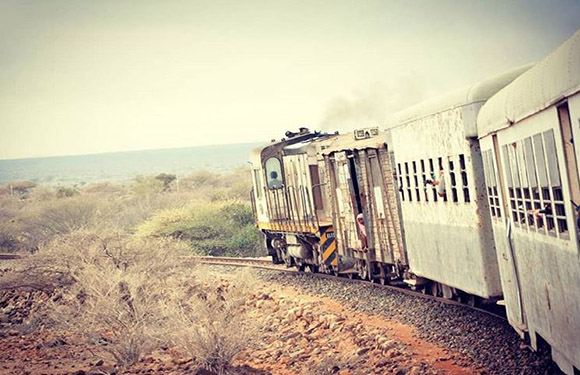 A train on a curved track in a dry, sparsely vegetated landscape emits smoke, with distant water visible.