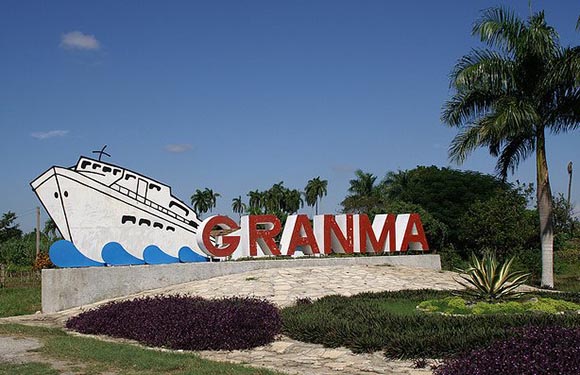 A monument with a large white boat sculpture labeled "GRANMA", set in a green, palm-filled landscape under a blue sky.