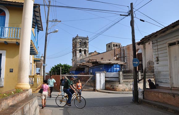 A street view in a town with people walking and cycling, old buildings, and overhead cables against a clear sky.