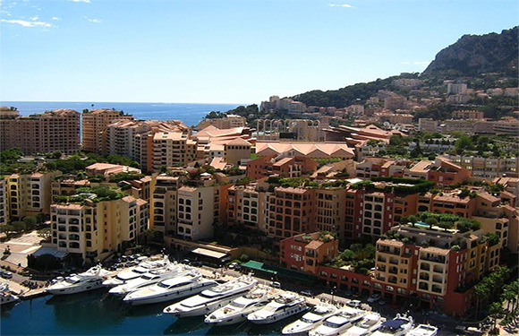 Aerial view of a coastal city with densely packed terracotta-roofed buildings, a marina with boats, and a mountain in the background.