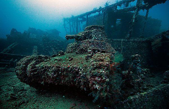 An underwater photograph showing a corroded tank covered in marine growth on the ocean floor, with a sunken structure visible in the background.