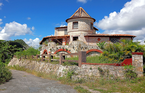 A stone castle-like building with a conical roof, surrounded by greenery under a partly cloudy sky.