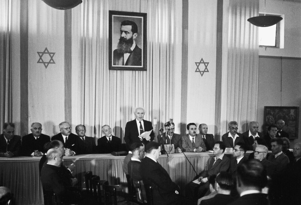 A black and white photo of a group of men seated in a formal setting with two large Star of David symbols on the wall, and a portrait hanging above.