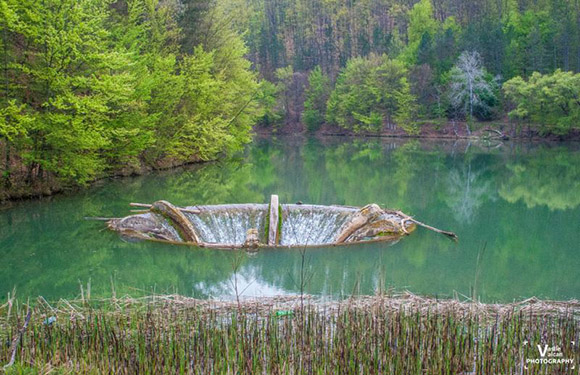 A small waterfall in a serene forest setting with lush green trees reflected in the calm water.