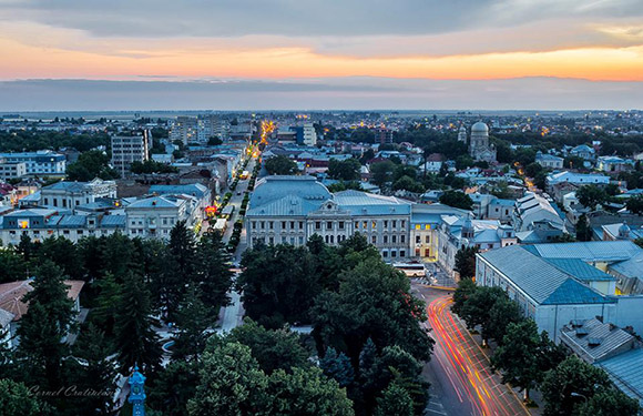 Aerial view of a city at dusk with illuminated streets and buildings, and a colorful sky in the background.