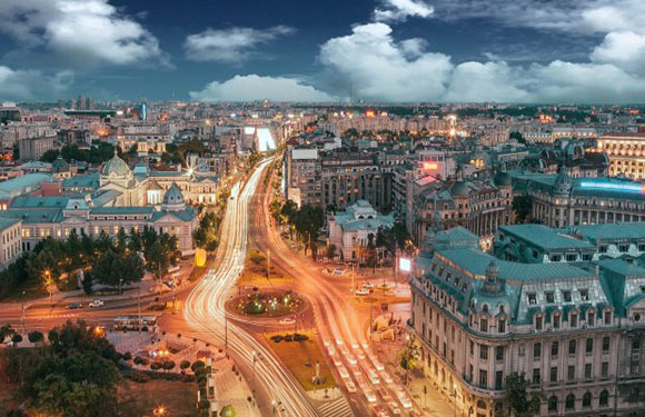 Aerial view of a city at dusk with illuminated streets and buildings, showcasing a blend of classical and modern architecture.