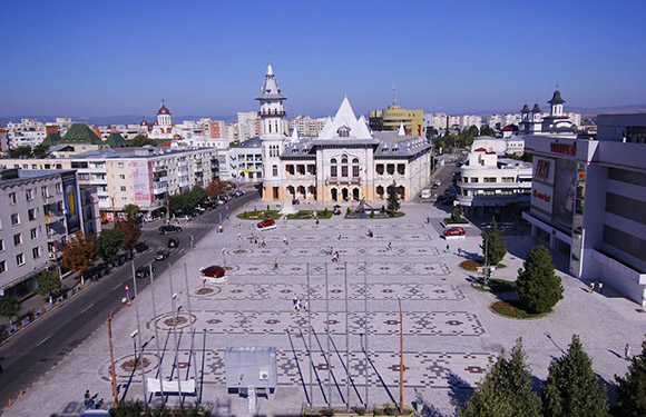 Aerial shot of a city square with patterned pavement, encircled by buildings and a church tower under a clear sky.