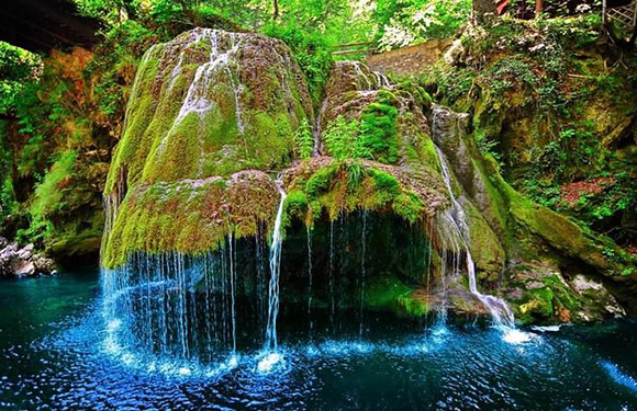 A small, vibrant waterfall with multiple streams cascading over moss-covered rocks into a clear blue pool surrounded by lush greenery.