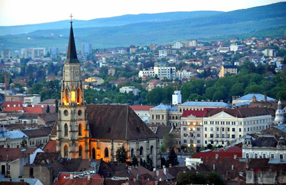 Aerial dusk view of a city with a tall, lit church spire, surrounded by buildings and distant hills.