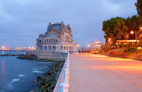 A twilight coastal promenade with a historic building, lit walkway with trees, and a calm sea.