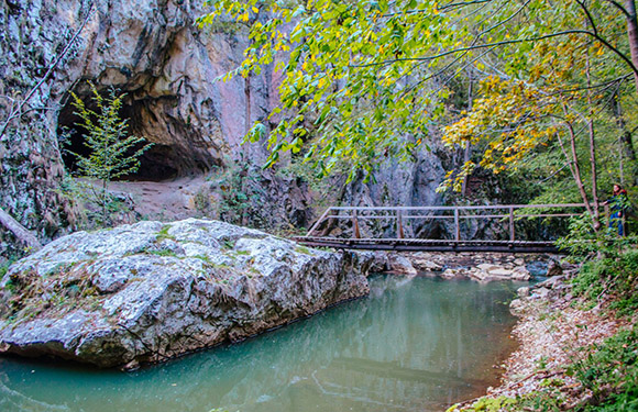 A serene nature scene with calm water, a rock, a metal bridge, autumn trees, and a partially hidden cave.