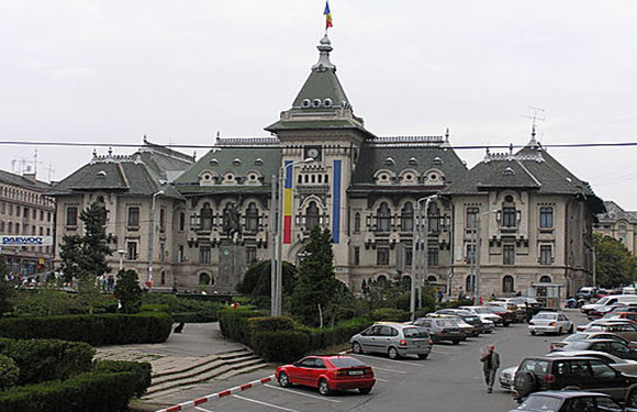 A photograph of a grand historic building with a central spire and flag, viewed from across a street with parked and moving cars.