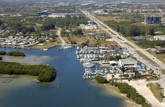 Aerial view of a coastal area with a marina, roads, buildings, and greenery.