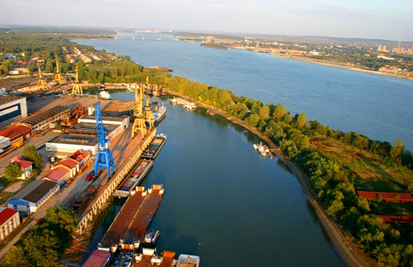 Aerial view of a river flowing through an industrial zone with docks and warehouses, town in the distance.