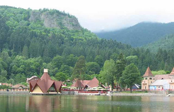 A scenic view of a lake with distinctive triangular-roofed buildings along the shore, backed by forested hills and a cloudy sky.