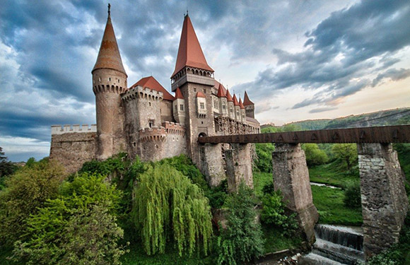 A picturesque view of a medieval castle with multiple spires and a stone bridge leading to it, set against a backdrop of greenery and a cloudy sky.