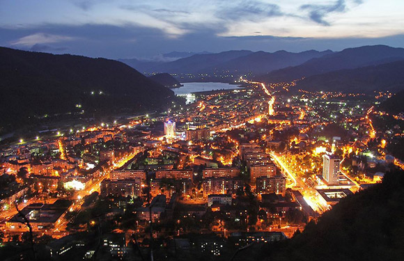 Aerial view of a city at night with lights illuminating streets and buildings, surrounded by hills and a body of water in the distance.