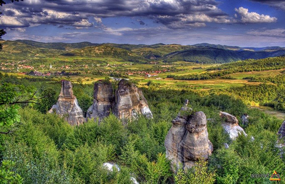 A picturesque scene with rock formations, greenery, rolling hills, clear sky, and a distant village.