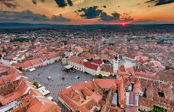 Aerial view of a historic town square at sunset with buildings arranged in a grid pattern, red rooftops, and a dramatic sky.