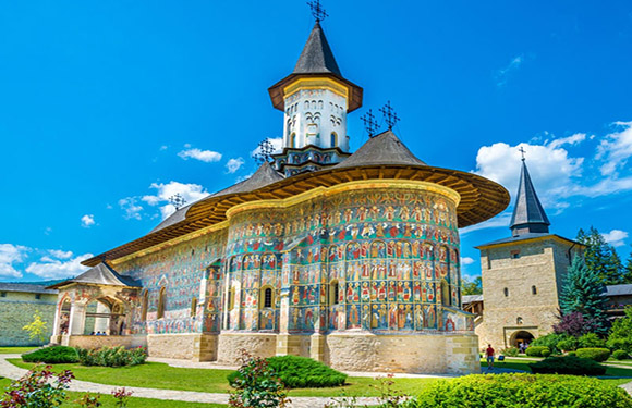 A vibrant monastery with exterior frescoes and a clock tower, set against a blue sky and lush greenery.