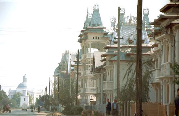 A street scene image with people, traditional buildings, ornate rooftops, trees, and utility poles.