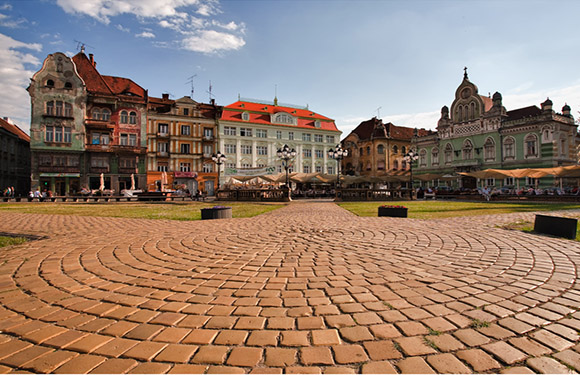 A cobblestone square with people and outdoor seating in front of colorful European-style buildings under a partly cloudy sky.