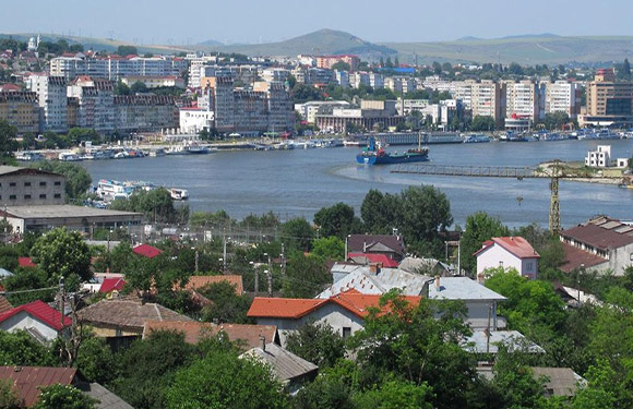 Aerial view of a coastal city with dense building clusters, a large body of water with boats, and hills in the background under a clear sky.