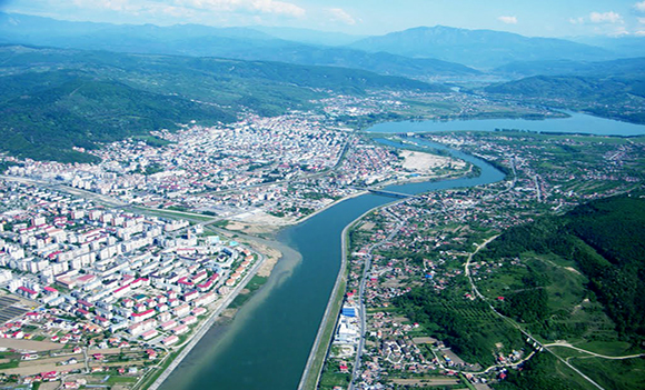 Aerial view of a city with a river dividing it, surrounded by green hills and mountains in the distance.