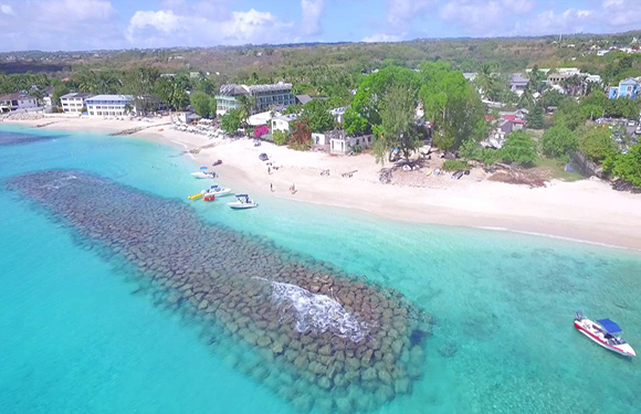 Aerial view of a tropical beach with clear turquoise waters, a breakwater, several boats, and beachfront buildings surrounded by greenery.