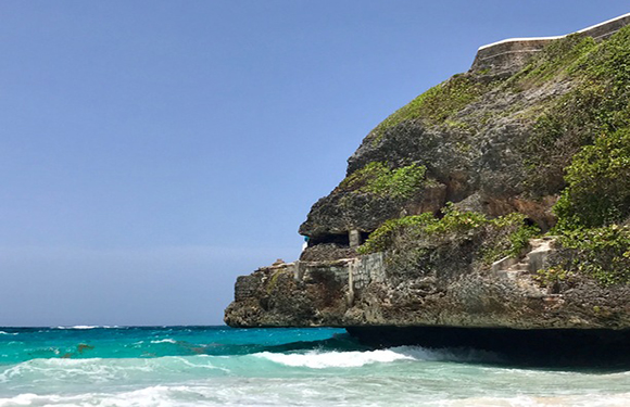 A coastal landscape with a large rocky cliff overhanging turquoise ocean waves under a clear blue sky.