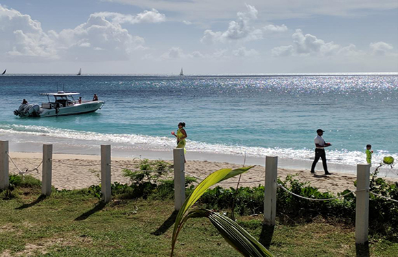 Two people stand on a beach near the water, with a boat nearby, under a bright sky. A fence and greenery are in the foreground.