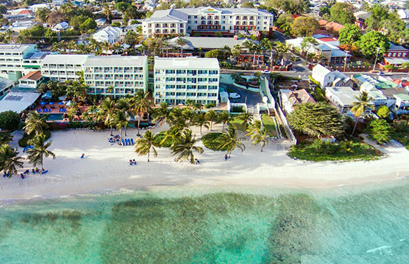 Aerial view of a tropical beachfront with buildings, palm trees, and people on the sand near the clear blue water.