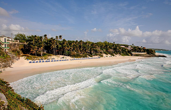 A tropical beach with white sand, turquoise water, palm trees, and a few beach chairs under the shade, with buildings on the coastline.