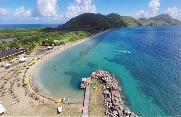 Aerial view of a tropical beach with a curved stone pier, clear blue waters, green hills in the background, and a few small boats on the shore.