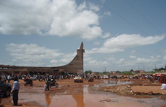 A busy outdoor scene with people, vehicles, a large monument, under a cloudy sky with puddles indicating recent rain.