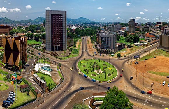 Aerial view of a cityscape with roads intersecting, surrounded by buildings and greenery under a partly cloudy sky.