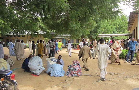 People gather outdoors in a dusty area with trees and a building, some standing, others sitting, with parked motorcycles nearby.