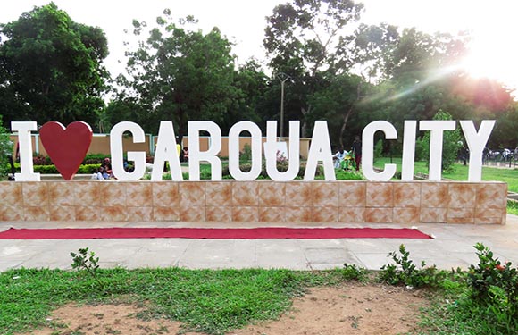 A sign in a sunny park reads "I LOVE GAROUA CITY," with a heart symbolizing love.