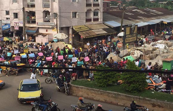 A busy street scene in a possible developing country, filled with crowds, stalls, motorcycles, and a yellow vehicle.