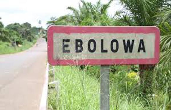 A weathered road sign with the text "EBOLOWA" on the side of a road with green vegetation in the background.