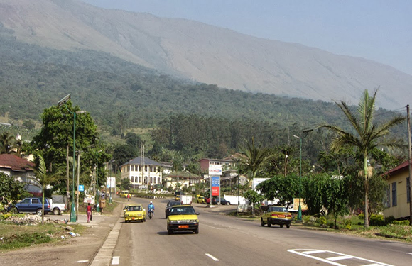 A road leading through a small town with buildings on either side, vehicles on the road, and a large mountain in the background under a clear sky.